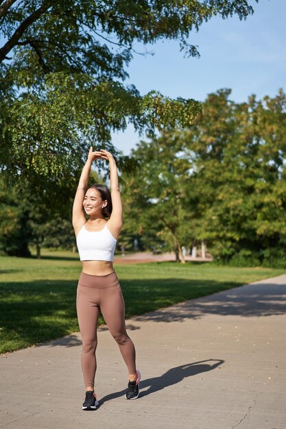 Chica asiática sonriente que se extiende después de un buen ejercicio en el parque escuchando música en auriculares inalámbricos jogg