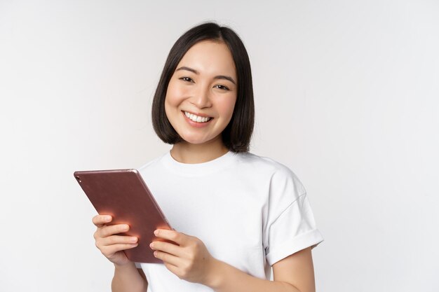 Chica asiática sonriente con tableta digital luciendo feliz y riendo posando en camiseta sobre fondo blanco