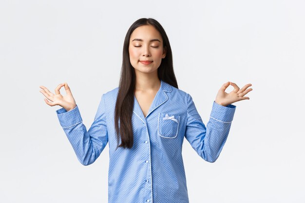 Chica asiática sonriente tranquila y feliz en pijama azul cierra los ojos, meditando antes de dormir o por la mañana, luciendo aliviada y en paz, practica la meditación del yoga sobre fondo blanco.