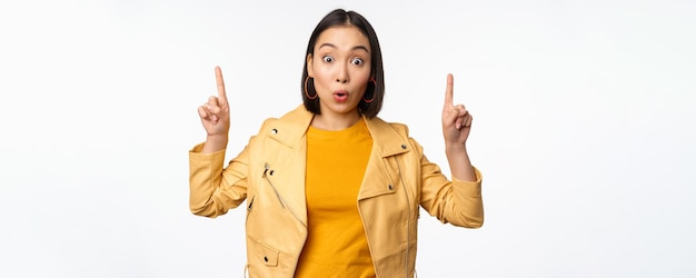 Chica asiática sorprendida señalando con el dedo hacia arriba expresa interés mostrando publicidad por delante demostrando banner promocional en la parte superior de pie contra fondo blanco