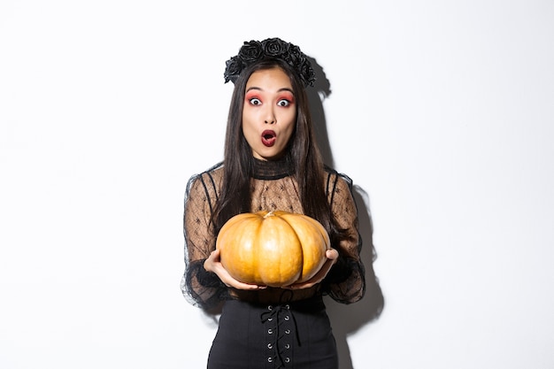 Foto gratuita chica asiática sorprendida en traje de bruja, sosteniendo calabaza y jadeando asombrada por la cámara, preparándose para la fiesta de halloween, de pie sobre fondo blanco.