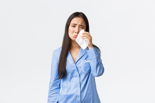 Chica asiática tonta angustiada en pijama azul con el corazón roto, quedarse en la cama de mal humor, limpiarse las lágrimas con un pañuelo, sollozar y llorar deprimida, afligida por el fondo blanco.