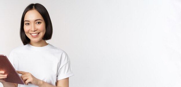 Chica asiática usando tableta digital trabajando con compras de gadgets en línea de pie sobre fondo blanco.