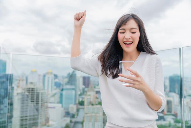 Chica asiática con vestido blanco sonrisa con dientes boca abierta emocionada y alegre con el resultado de la comprobación de la pantalla del teléfono inteligente que muestra el fondo del paisaje urbano del edificio de capital