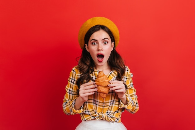 Chica asombrada con cabello ondulado con delicioso bollo. Mujer de ojos verdes vestida de amarillo quiere comer croissant.