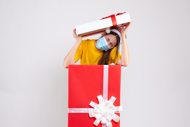 Chica asustada con gorro de Papá Noel poniendo la tapa de la caja en la cabeza de pie detrás de un gran regalo de Navidad en blanco