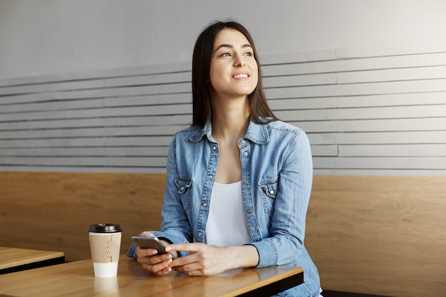 Chica atractiva alegre con el pelo oscuro sentada en la cafetería, bebe café y conversa con un amigo en el teléfono inteligente y luego gira la cabeza para ver al novio a través de la ventana.