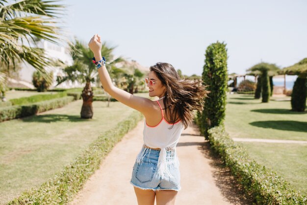 Chica atractiva en buena forma divertida bailando con el pelo ondeando y riendo, disfrutando de las vacaciones de verano en un país exótico. Retrato de mujer joven morena sonriente en gafas de sol de moda corriendo en el parque.