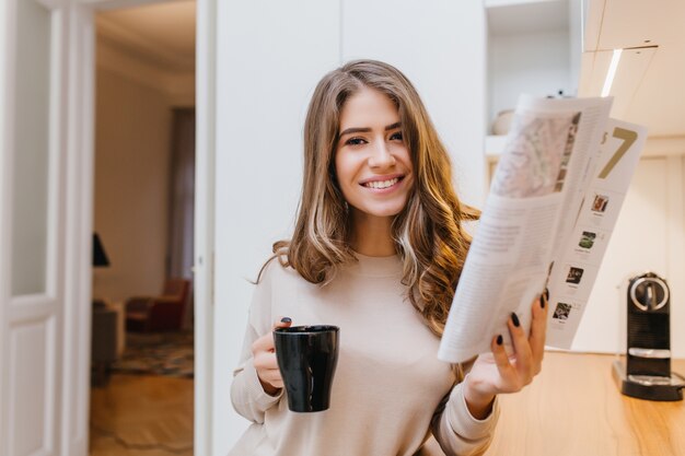 Chica atractiva con cabello castaño claro sosteniendo una revista y riendo en su habitación