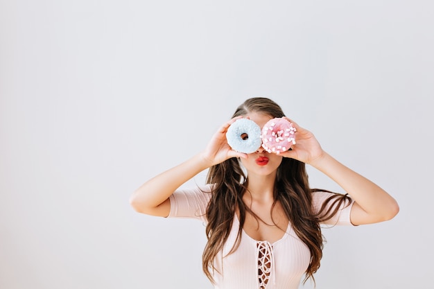 Chica atractiva con cabello largo morena sosteniendo donuts coloridos contra sus ojos. Mujer joven alegre con labios rojos divirtiéndose con dulces, delicioso. Vida luminosa. expresiones, concepto de dieta.