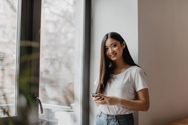 Chica atractiva con cabello largo sosteniendo teléfono inteligente mirando a la cámara y posando junto a la ventana