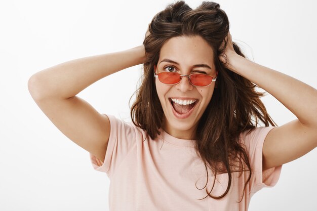 Chica atractiva descarada con gafas de sol posando contra la pared blanca