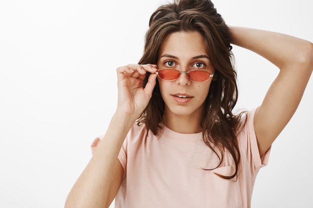 Chica atractiva descarada con gafas de sol posando contra la pared blanca