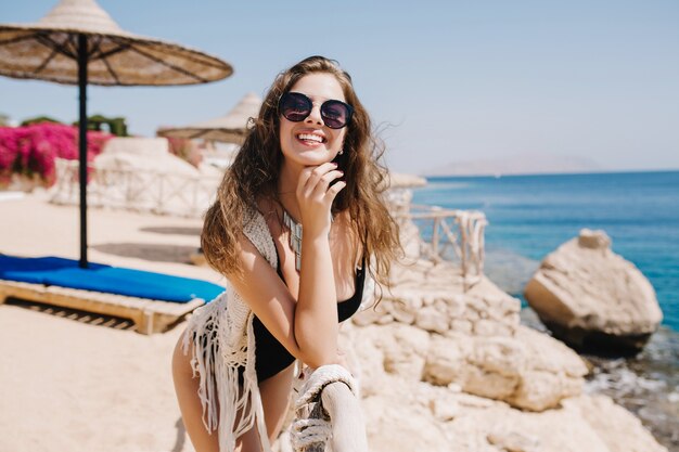 Chica atractiva en elegantes gafas de sol riendo, mientras posa en la orilla del mar con horizonte. Mujer joven alegre con cabello castaño claro y sonrisa increíble descansando en la playa del océano