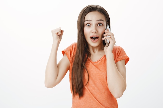 Chica atractiva emocionada y sorprendida hablando por teléfono móvil y animando, ganando y saltando de la felicidad