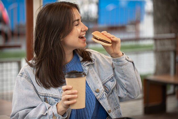 Chica atractiva en estilo casual come una hamburguesa con café sentado en la terraza de verano