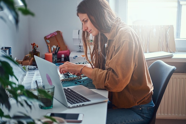 Foto gratuita una chica atractiva está haciendo collares con cuentas y viendo instrucciones en la computadora.