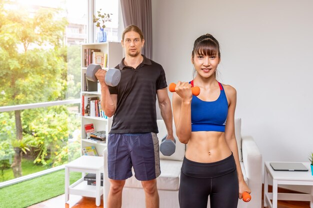 Chica atractiva haciendo ejercicios de peso con mancuernas con entrenador personal en casa mirando a la cámara