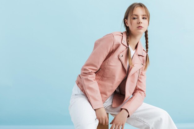 Chica atractiva joven con dos trenzas en chaqueta de cuero rosa y jeans blancos mirando cuidadosamente en cámara mientras se apoya en las rodillas sobre fondo azul.