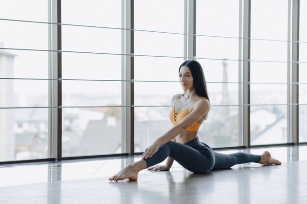 Chica atractiva joven haciendo ejercicios de fitness con yoga en el suelo contra la superficie de las ventanas panorámicas