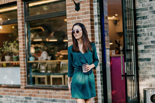 Chica atractiva de moda en vestido verde de verano y gafas de sol está caminando en el parque de la ciudad con café para llevar y pasar el fin de semana al aire libre