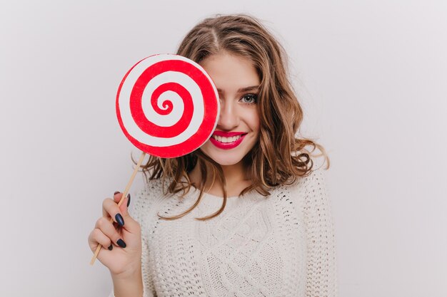 Chica atractiva con ojos grises y labios brillantes está sonriendo, cubriendo parte de su rostro con piruleta