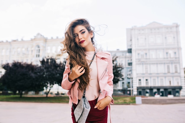 Chica atractiva con peinado largo en pantalones vinosos está posando en la ciudad.