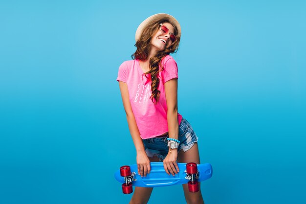 Chica atractiva con pelo largo y rizado con sombrero posando sobre fondo azul en estudio. Lleva pantalones cortos, camiseta rosa, gafas de sol rosas. Ella sostiene una patineta azul.