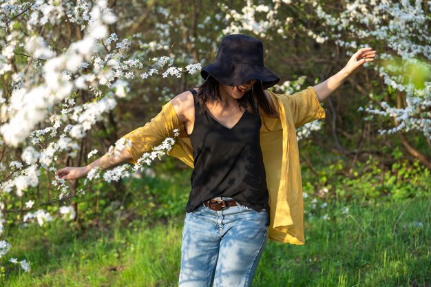 Una chica atractiva con un sombrero entre árboles en flor disfruta del olor de las flores de primavera.