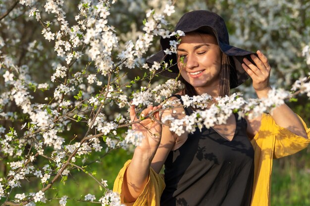 Una chica atractiva con un sombrero entre árboles en flor disfruta del olor de las flores de primavera