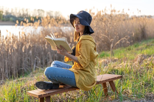 Chica atractiva con un sombrero lee un libro en la naturaleza al atardecer.