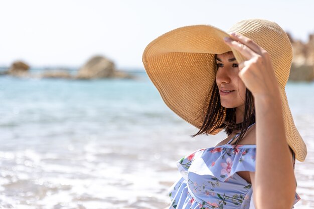 Chica atractiva con un sombrero oculta su rostro del sol, sentada en la orilla del mar.