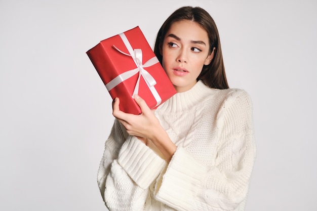 Chica atractiva en suéter acogedor con caja de regalo mirando cuidadosamente hacia otro lado sobre fondo blanco aislado