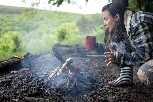 Una chica atractiva con una taza en la mano se calienta cerca de un fuego en el bosque.