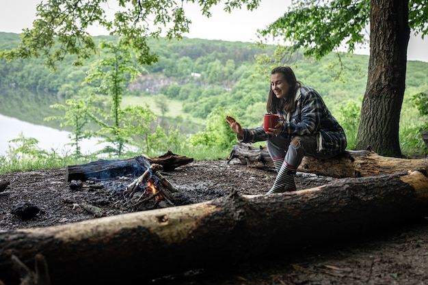 Una chica atractiva con una taza en la mano se sienta en un tronco y se calienta cerca de un fuego en el bosque.