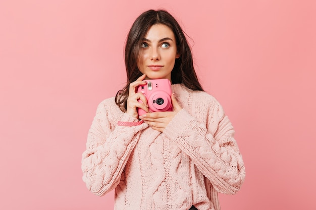 Chica atractiva toma fotografías en la cámara instax. Señora en suéter rosa busca sobre fondo aislado.