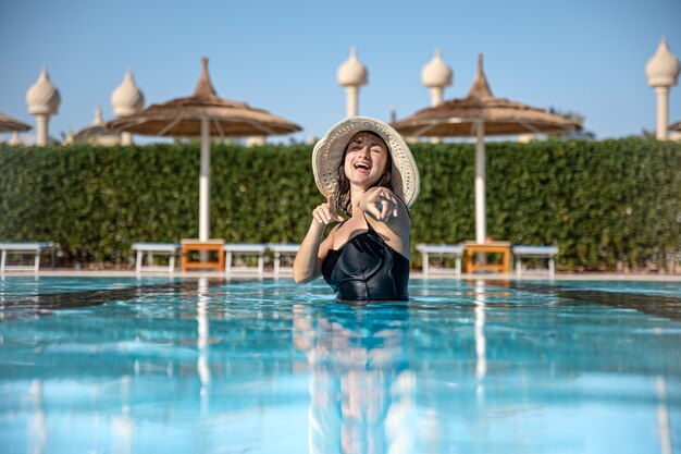 Chica atractiva en traje de baño negro y sombrero se baña en la piscina. El concepto de vacaciones y recreación en un país cálido.