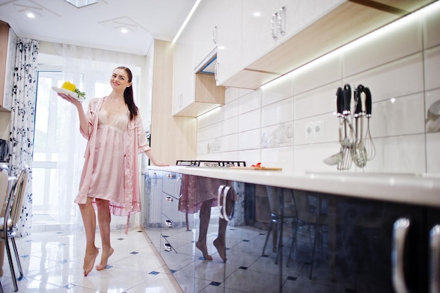 Chica atractiva en túnica rosa posando con un plato de ensalada en la cocina