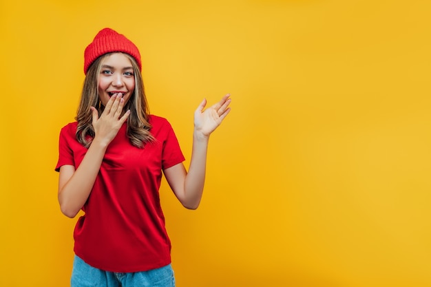 Chica atractiva vestida con ropa roja sobre fondo amarillo muestra su mano en su lugar para el texto y sorprendida, con una sonrisa