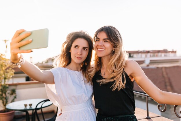 Chica atractiva con vestido blanco y accesorios de moda tomando selfie con mejor amigo en la cafetería al aire libre en la mañana