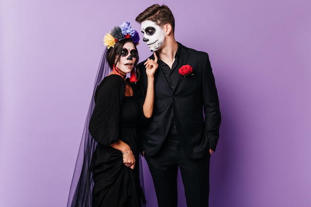 Chica atractiva zombie en guirnalda colorida con novio. pareja caucásica alegre en disfraces de muertos posando en halloween sobre fondo púrpura.