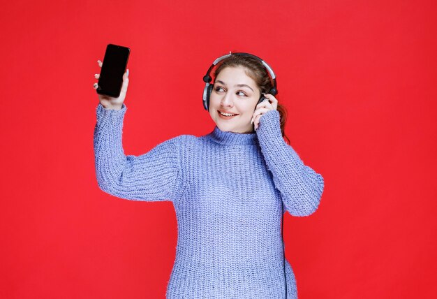 Chica con auriculares demostrando su teléfono inteligente y sintiéndose feliz.