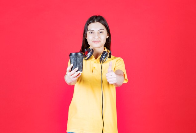 Chica con auriculares sosteniendo una taza de bebida desechable negra y mostrando un signo de mano positivo