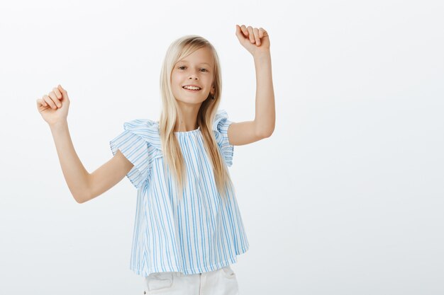 Chica bailando en la fiesta de amigos, divirtiéndose. Retrato interior de una niña alegre alegre positiva con cabello rubio, levantando las manos y haciendo movimientos de baile con una sonrisa feliz