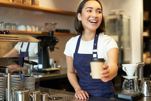 Foto gratuita una chica barista asiática sonriente que da una taza de café para llevar prepara el pedido para llevar al huésped en la cafetería usando