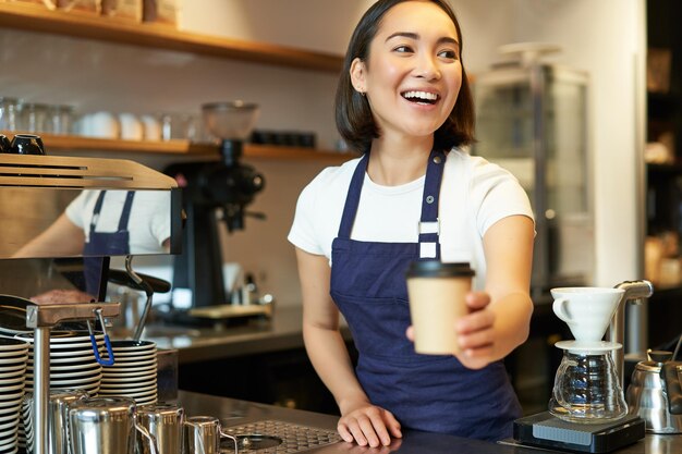 Una chica barista asiática sonriente que da una taza de café para llevar prepara el pedido para llevar al huésped en la cafetería usando