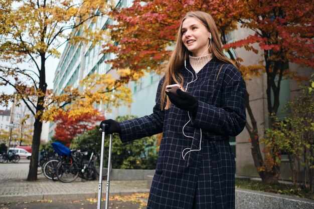 Chica bastante alegre en auriculares felizmente escuchando música en el teléfono celular esperando en la calle con maleta