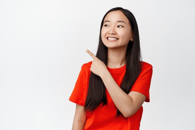 Chica bastante asiática sonriendo señalando y mirando el espacio vacío de la esquina superior izquierda que muestra el logotipo o el anuncio de venta con expresión de cara satisfecha fondo blanco
