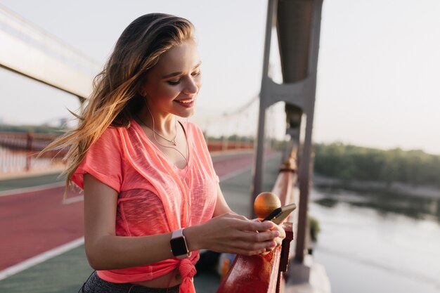 Chica bastante bien formada en mensaje de texto de reloj de pulsera. Riendo a mujer caucásica con teléfono después del entrenamiento.