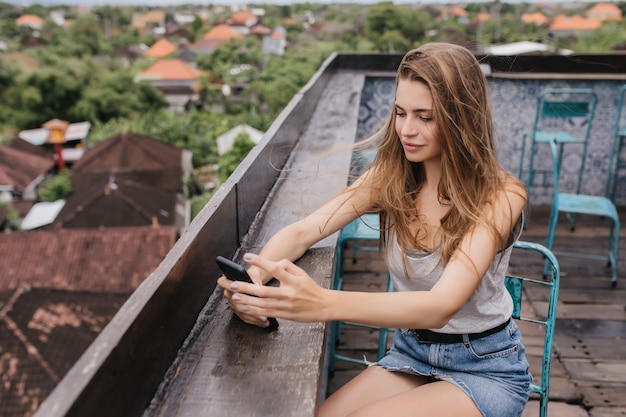 Chica bastante caucásica en falda de mezclilla sentada en el techo con smartphone. Retrato al aire libre de una dama de cabello castaño escalofriante en la calle.